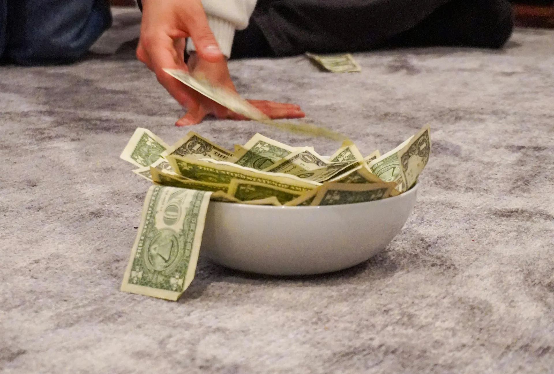 Person dropping dollar bills into a bowl overflowing with money, on a gray carpet.